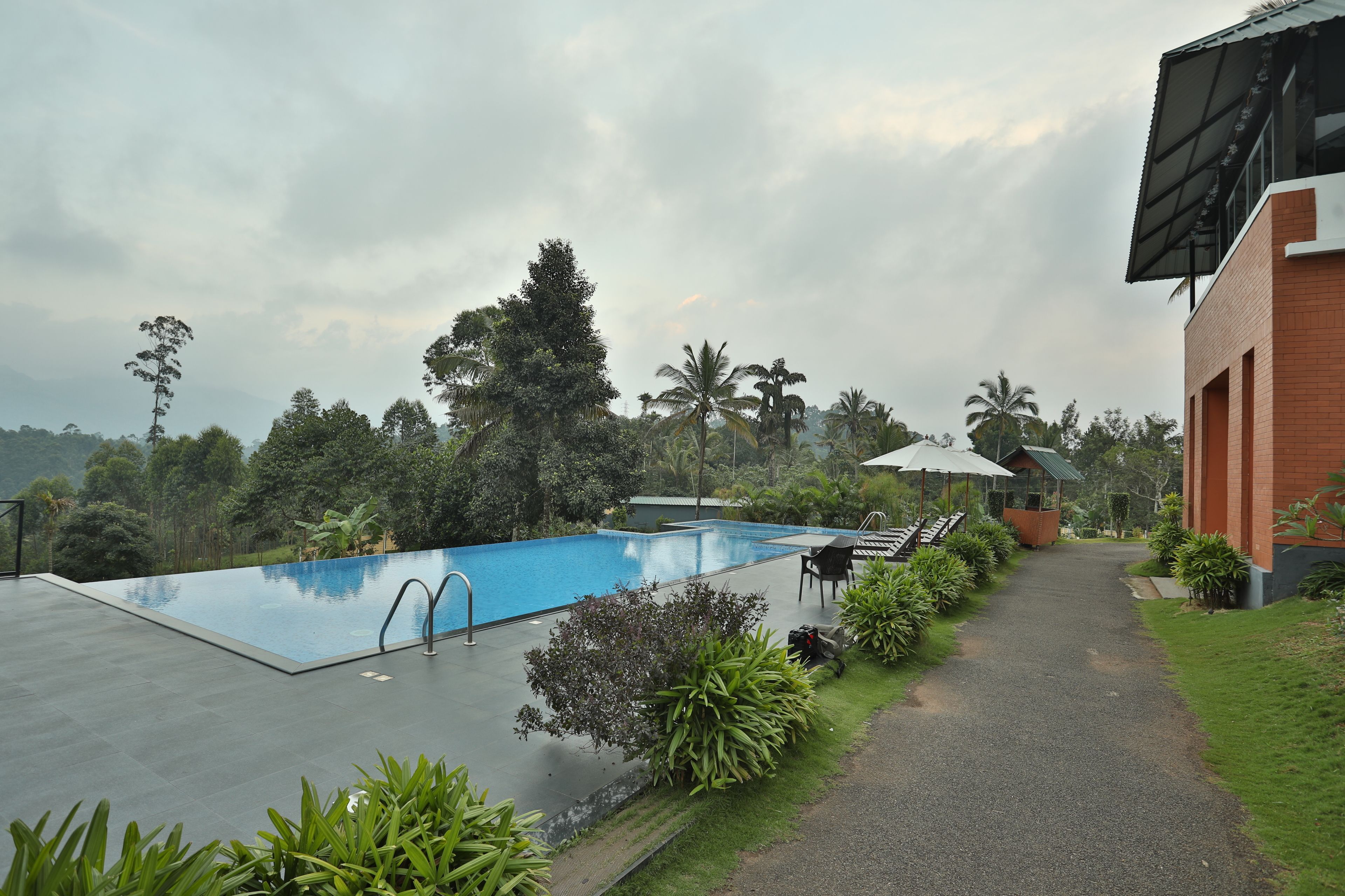A swimming pool with a clear blue water, surrounded by lush greenery and a pathway leading to a building with a sloped roof.