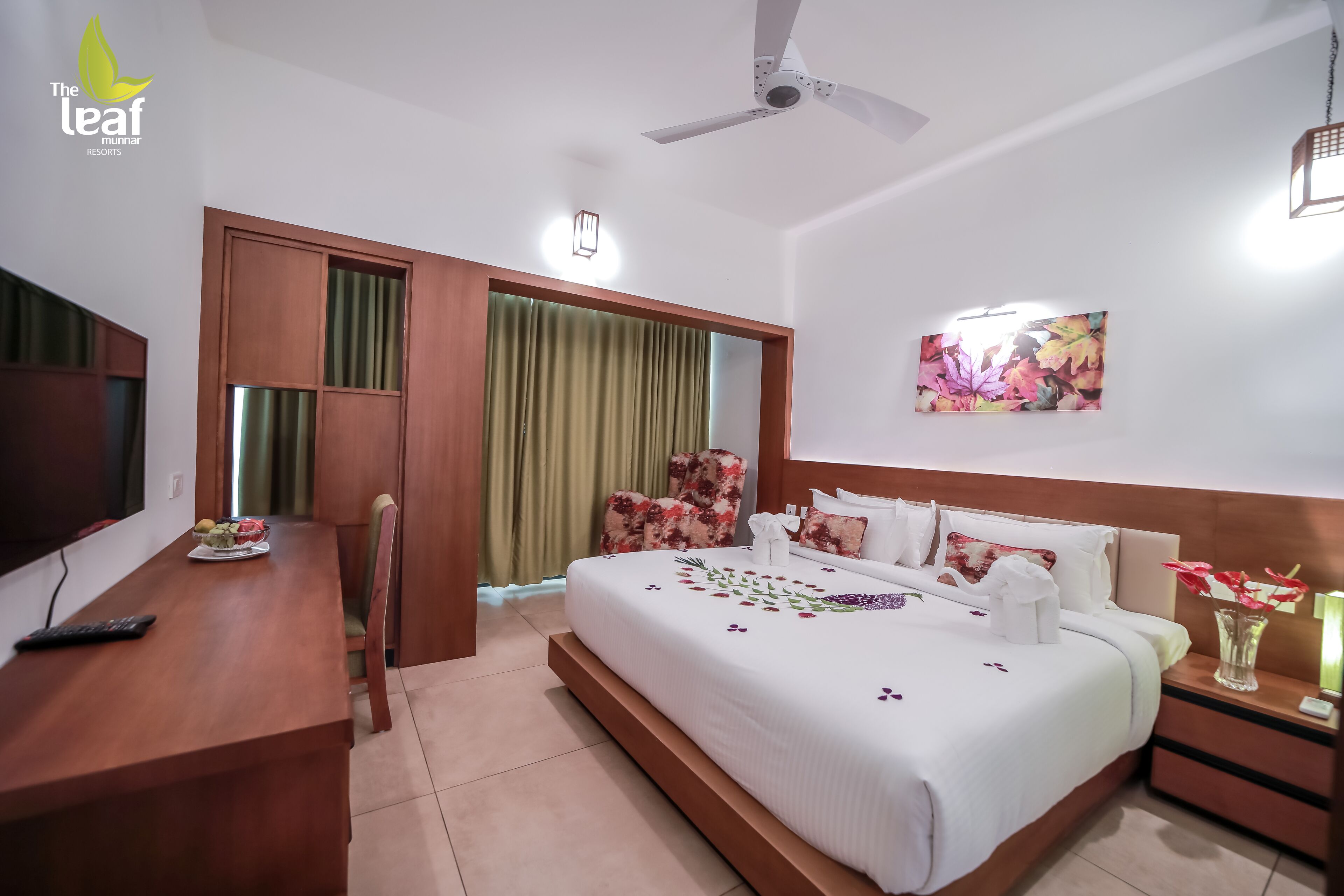 A hotel room with a bed, a desk, a chair, a TV, a ceiling fan, and a painting.