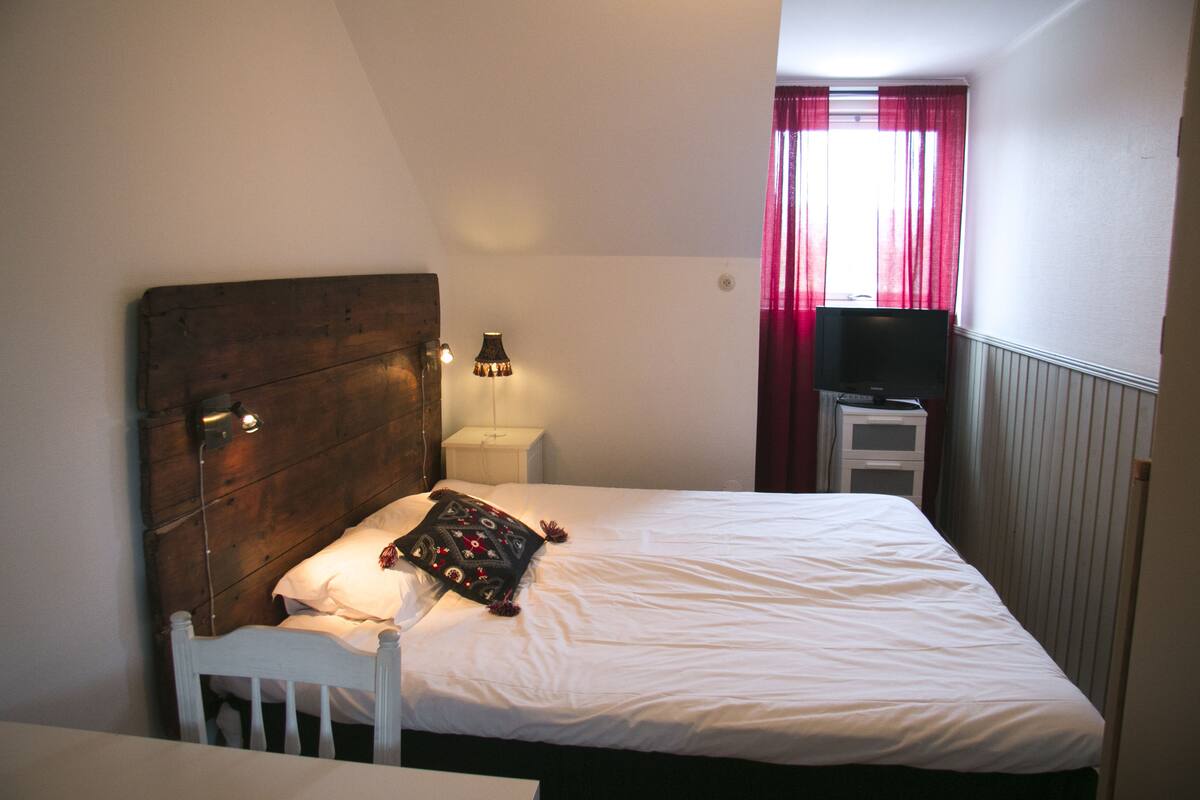 A bedroom with a wooden headboard, a white bed, a nightstand with a lamp, a TV on a stand, and a window with red curtains.