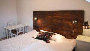 Un dormitorio con cabecera de madera, una cama individual con ropa de cama blanca, una mesita de noche con lámpara y una silla blanca.