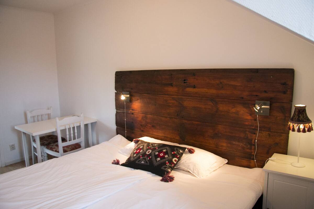 A bedroom with a wooden headboard, a single bed with white bedding, a nightstand with a lamp, and a white chair.