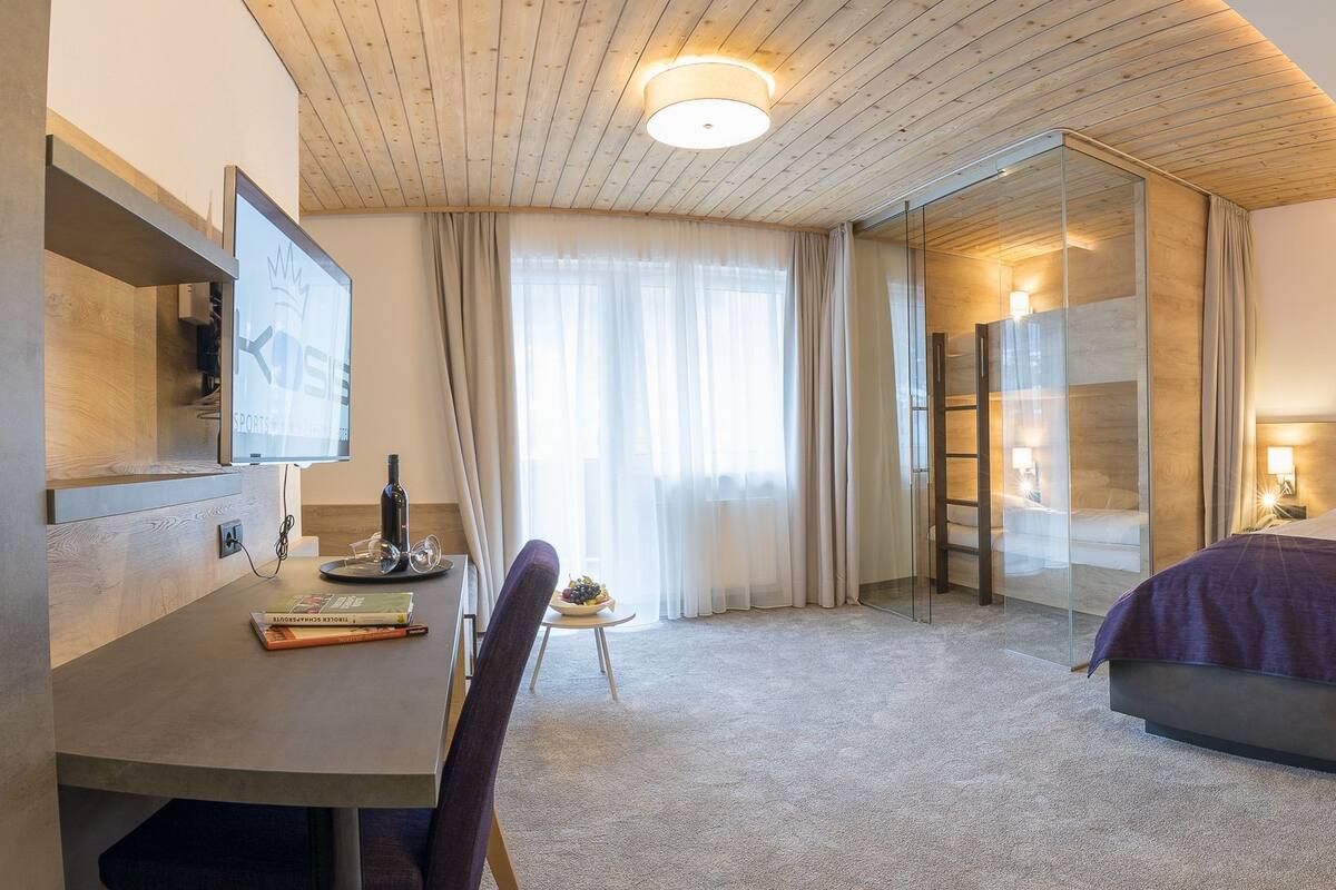 Moderni hotellihuone, jossa on suuri sänky, työpöytä tuolilla, televisio ja lasiseinä.