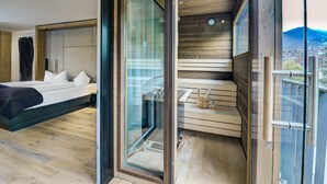 A modern bedroom with a bed, a glass door leading to a balcony, and a wooden sauna area. - KOSIS Sports Lifestyle Hotel (Fuegen)