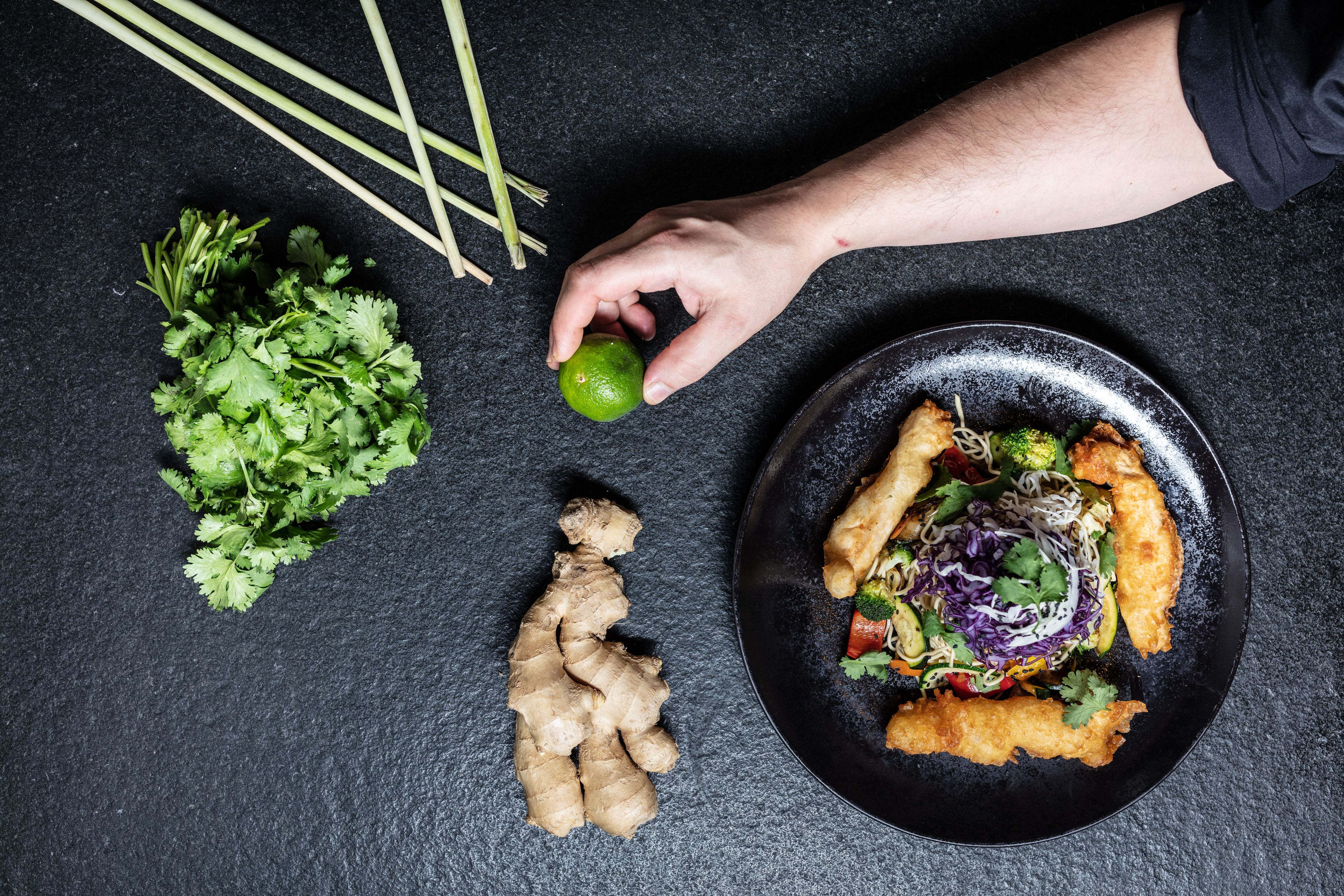 Une assiette de nourriture contenant des légumes et des herbes aromatiques, un citron vert et du gingembre, le tout posé sur une surface sombre.