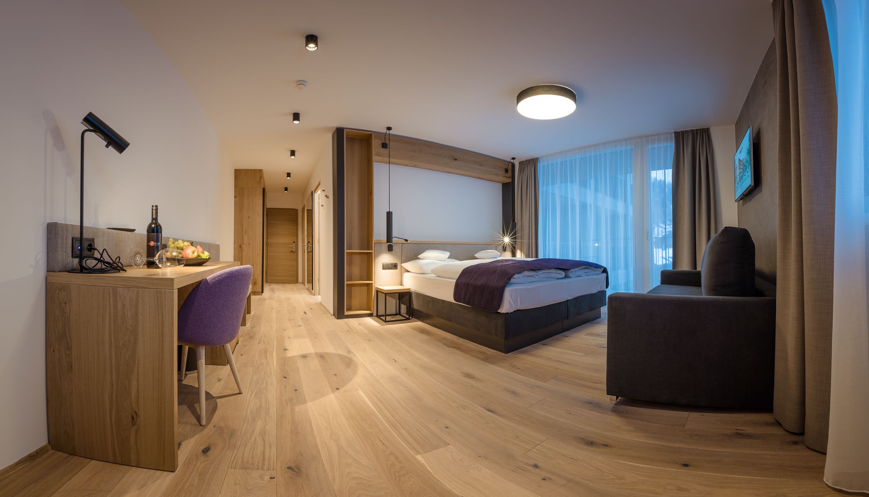 A modern hotel room with a wooden desk, a purple chair, a bed with a headboard, a TV, and a large window with curtains.