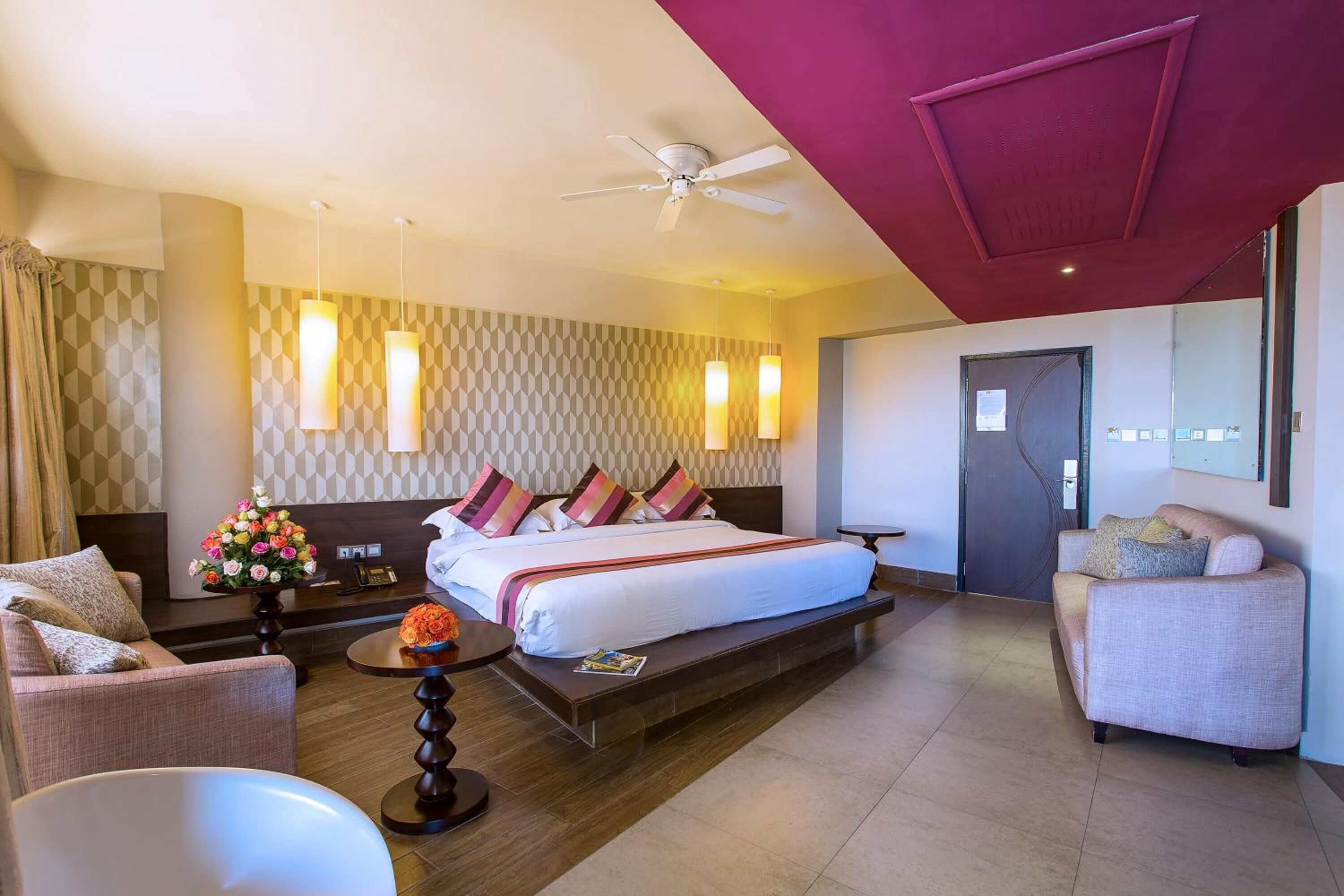 A hotel room with a large bed, a sofa, a chair, a small table with a vase of flowers, and a ceiling fan.