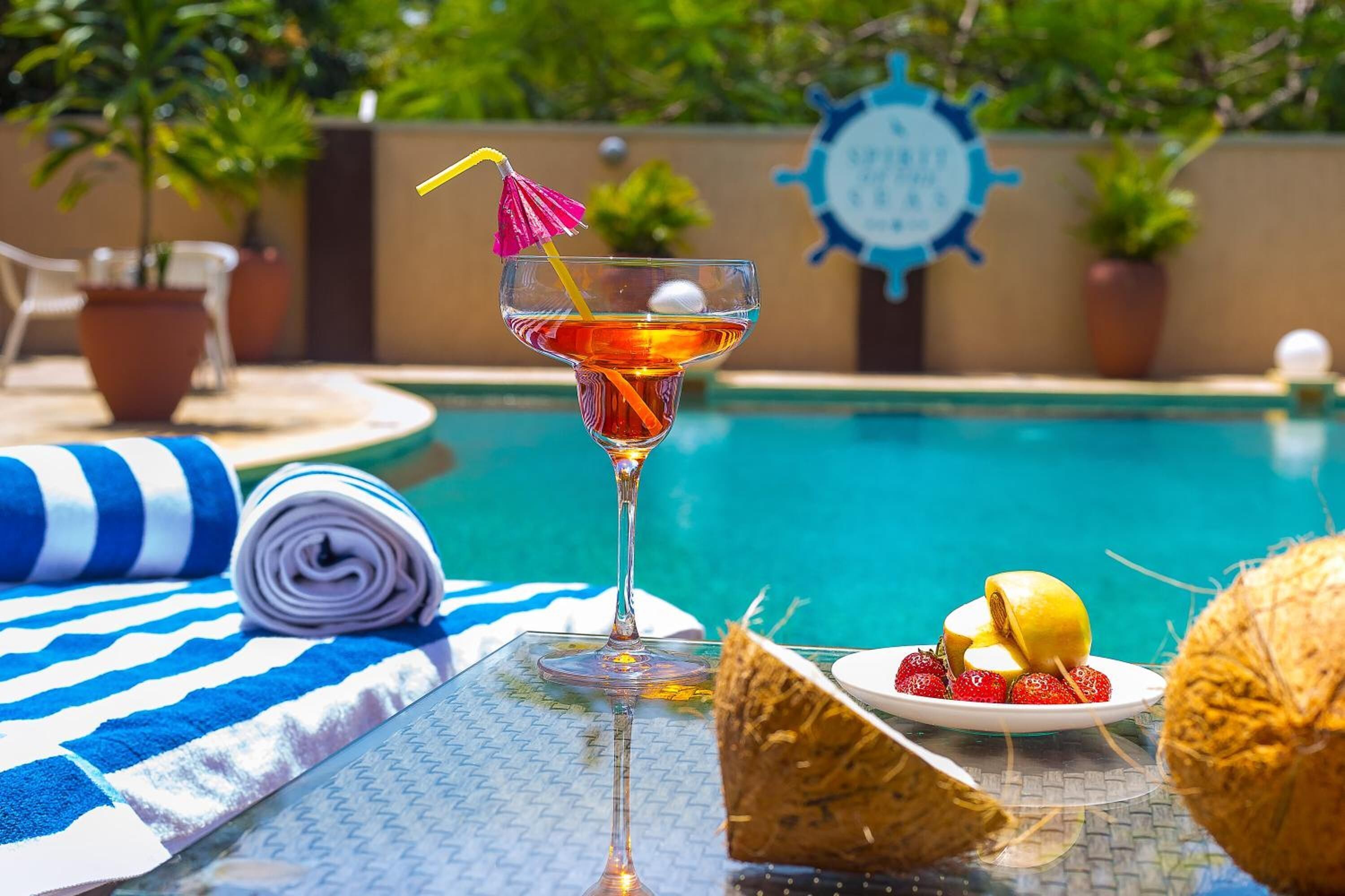 Ambiente à beira da piscina com um copo de cocktail, um prato de morangos, uma toalha enrolada e um guarda-sol de palha.