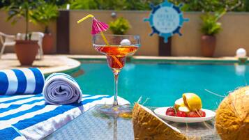 Ambiente à beira da piscina com um copo de cocktail, um prato de morangos, uma toalha enrolada e um guarda-sol de palha.