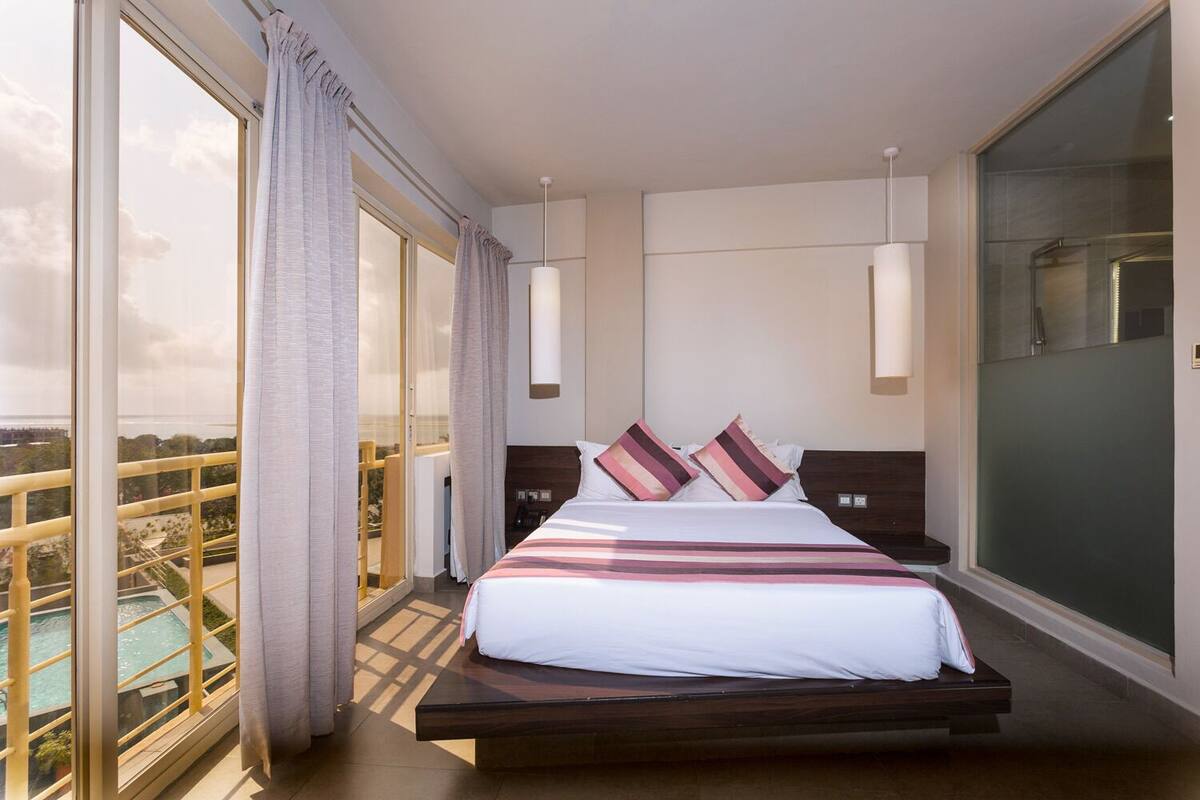 Quarto de hotel moderno com uma cama grande, vista para uma piscina e varanda com guarda-corpo de vidro.