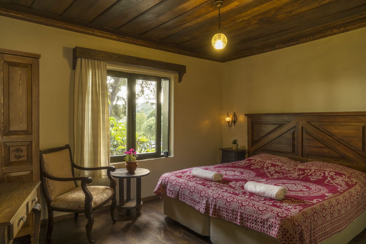 A bedroom with a wooden headboard, a bed with a patterned bedspread, a chair, a small table with a potted plant, and a window with curtains.