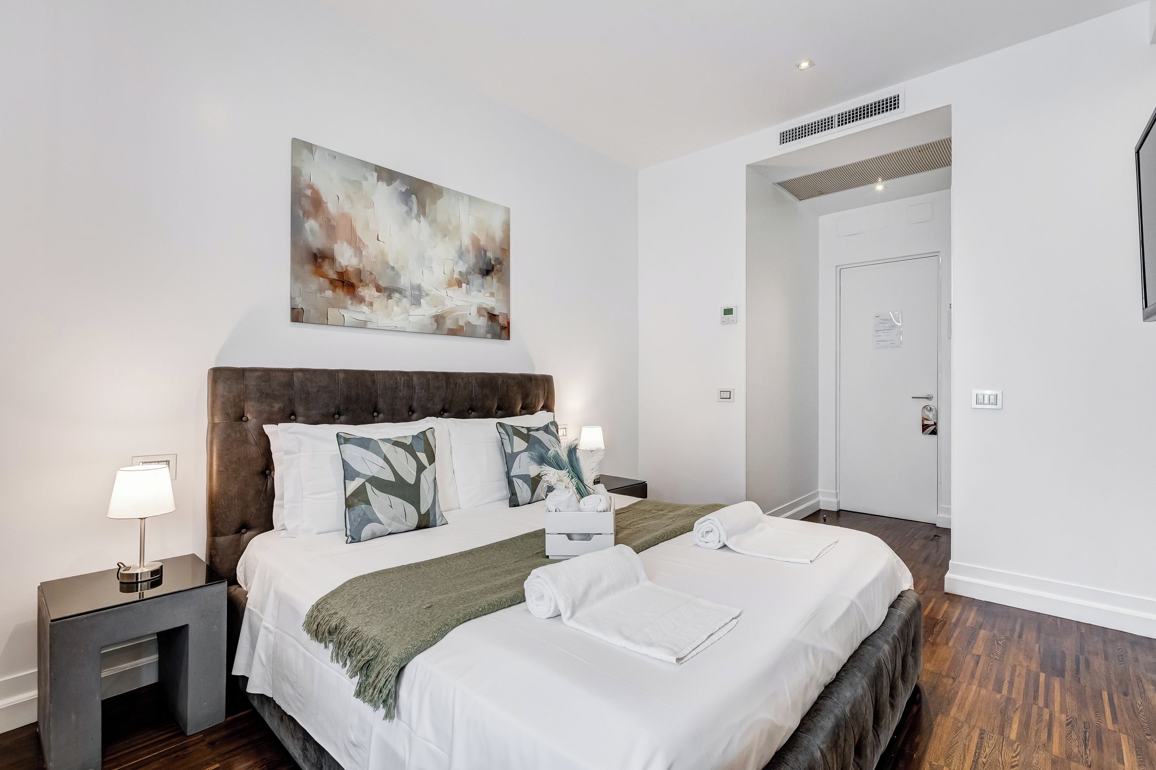 A hotel room with a large bed, two bedside tables, a wall-mounted abstract painting, and a door leading to another room.