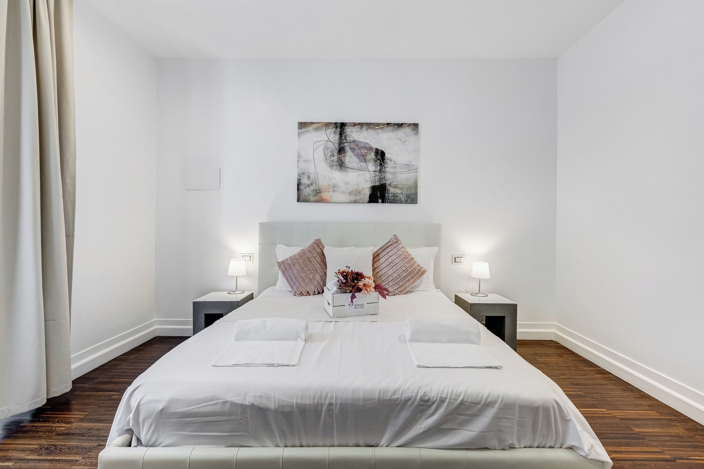 A hotel room with a large bed, two bedside tables, a nightstand, a wall-mounted artwork, and a window with curtains.