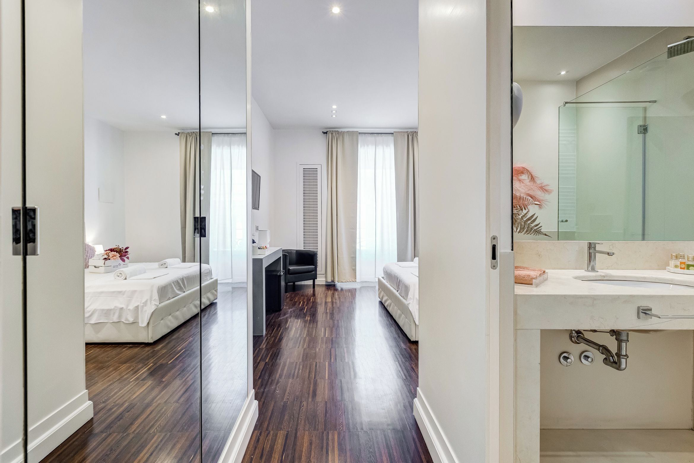 Habitación de hotel con dos camas, un escritorio, una silla, un espejo grande y baño con lavamanos y ducha.