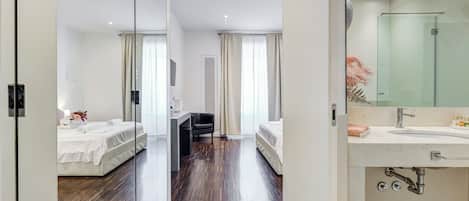 Habitación de hotel con dos camas, un escritorio, una silla, un espejo grande y baño con lavamanos y ducha.