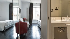 Una habitación de hotel con una cama, una silla roja, una mesita con un bolso y un baño con un lavabo y un espejo.
