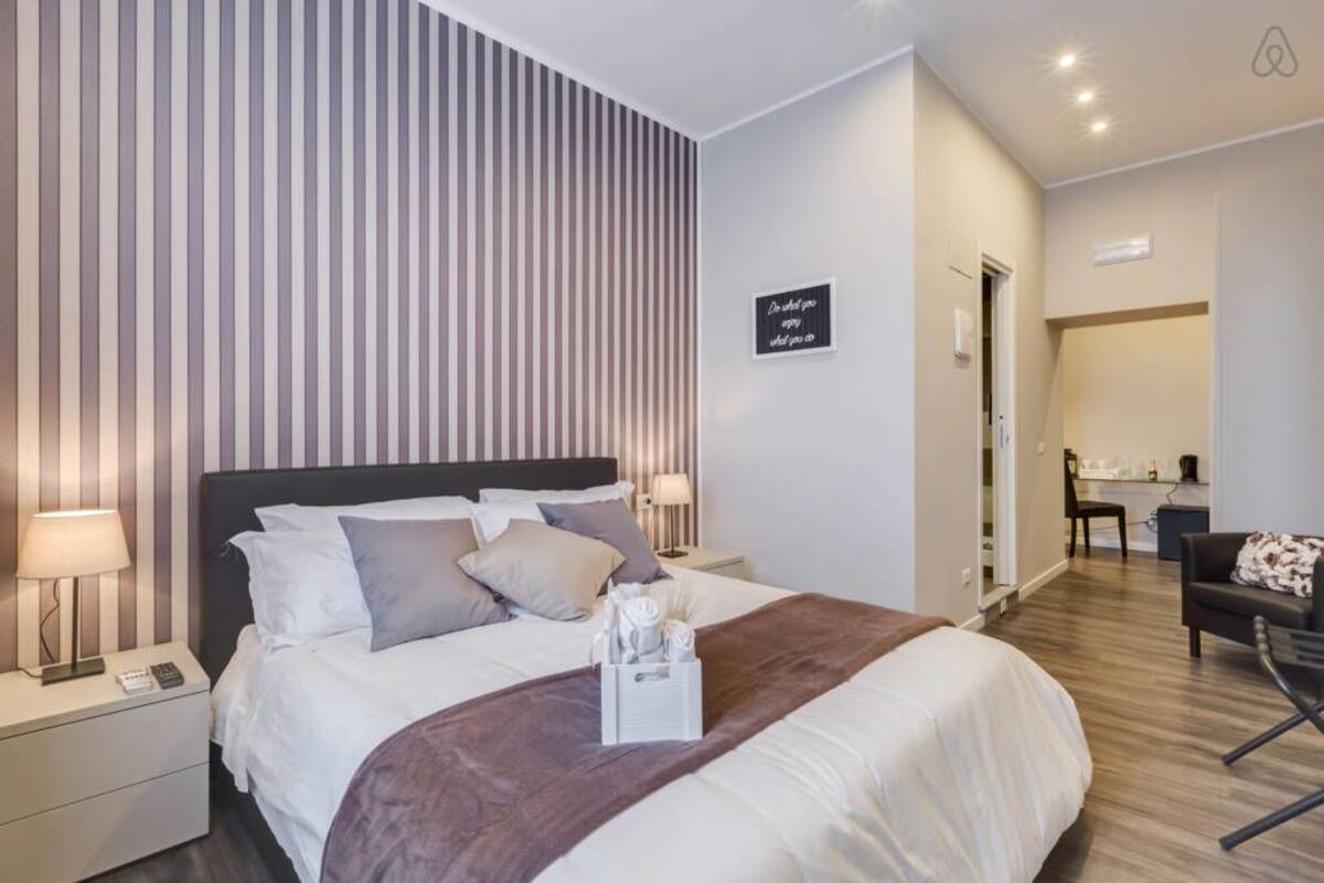 A modern hotel room with a large bed, a bedside table with a lamp, a wall-mounted sign, and a view into the dining area.