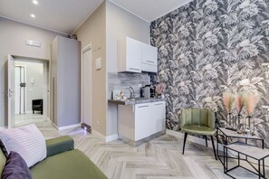 A modern living room with a sofa, a small table, a green armchair, and a kitchen area with white cabinets and a tiled backsplash. - Hanky Suite (Rome)