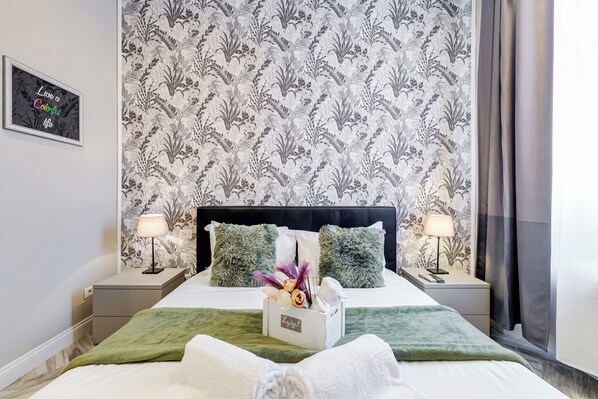 Une chambre d’hôtel comprenant un lit, deux tables de chevet, une petite table, un tableau accroché au mur et une fenêtre avec des rideaux.