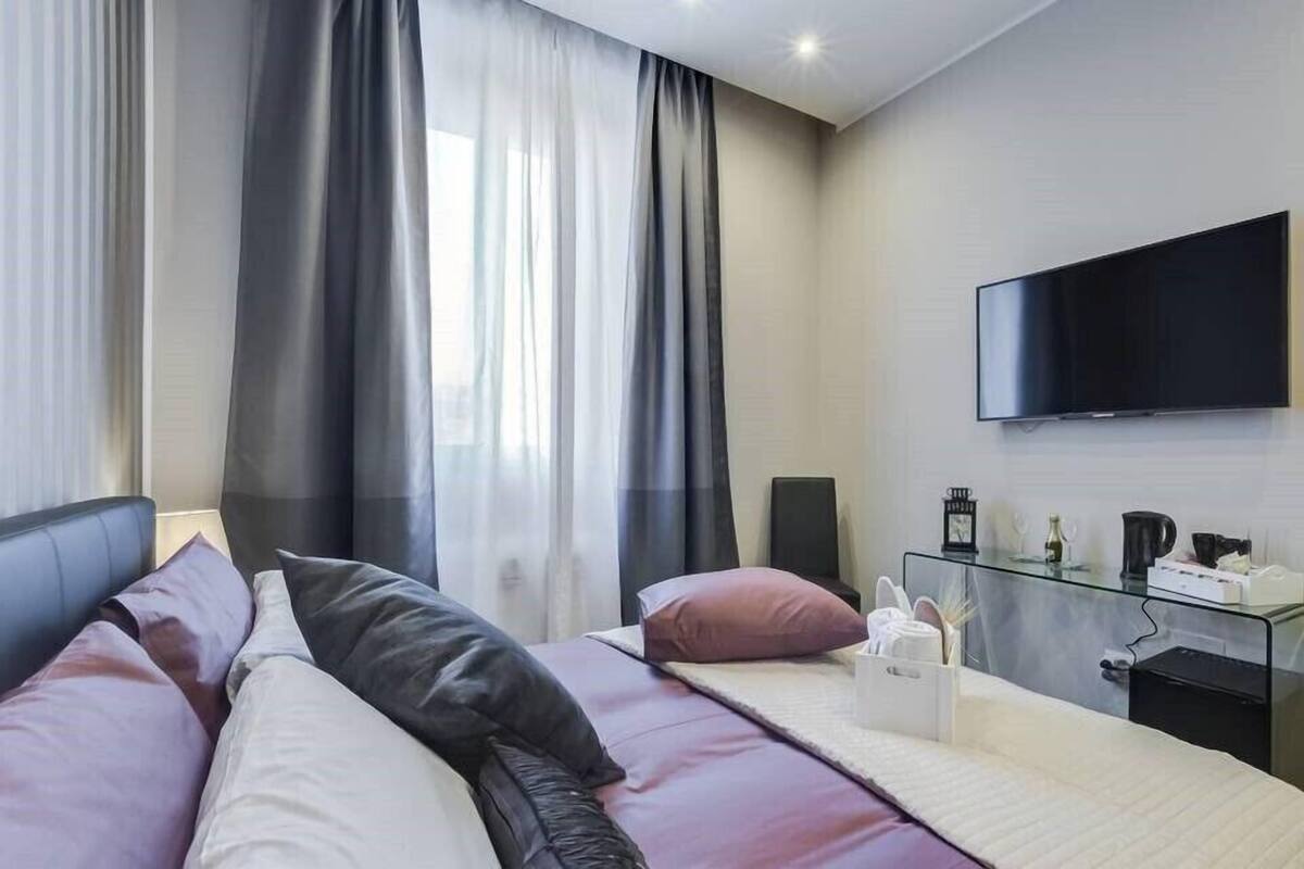 A modern hotel room with a bed, curtains, a flat-screen TV, and a glass side table with a coffee maker.