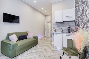 A modern living space with a sofa, a flat-screen TV, a kitchen with white cabinets, and a herringbone-patterned floor. - Hanky Suite (Rome)