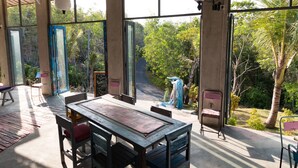 Um quarto espaçoso com uma mesa grande e cadeiras, janelas amplas com vista para vegetação exuberante.
