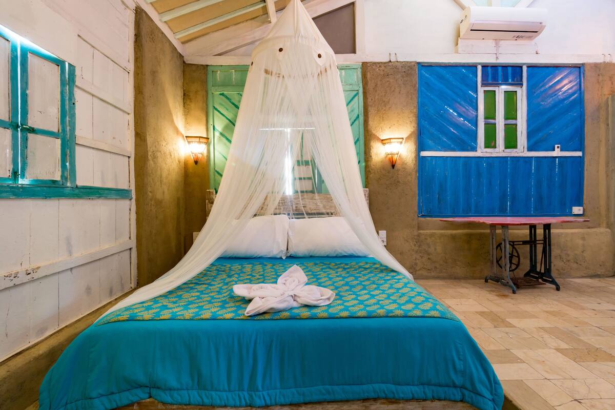A room with a bed under a white canopy, a blue and yellow patterned bedspread, a white towel on the bed, a table with a red and black tablecloth, and a blue window with green curtains.