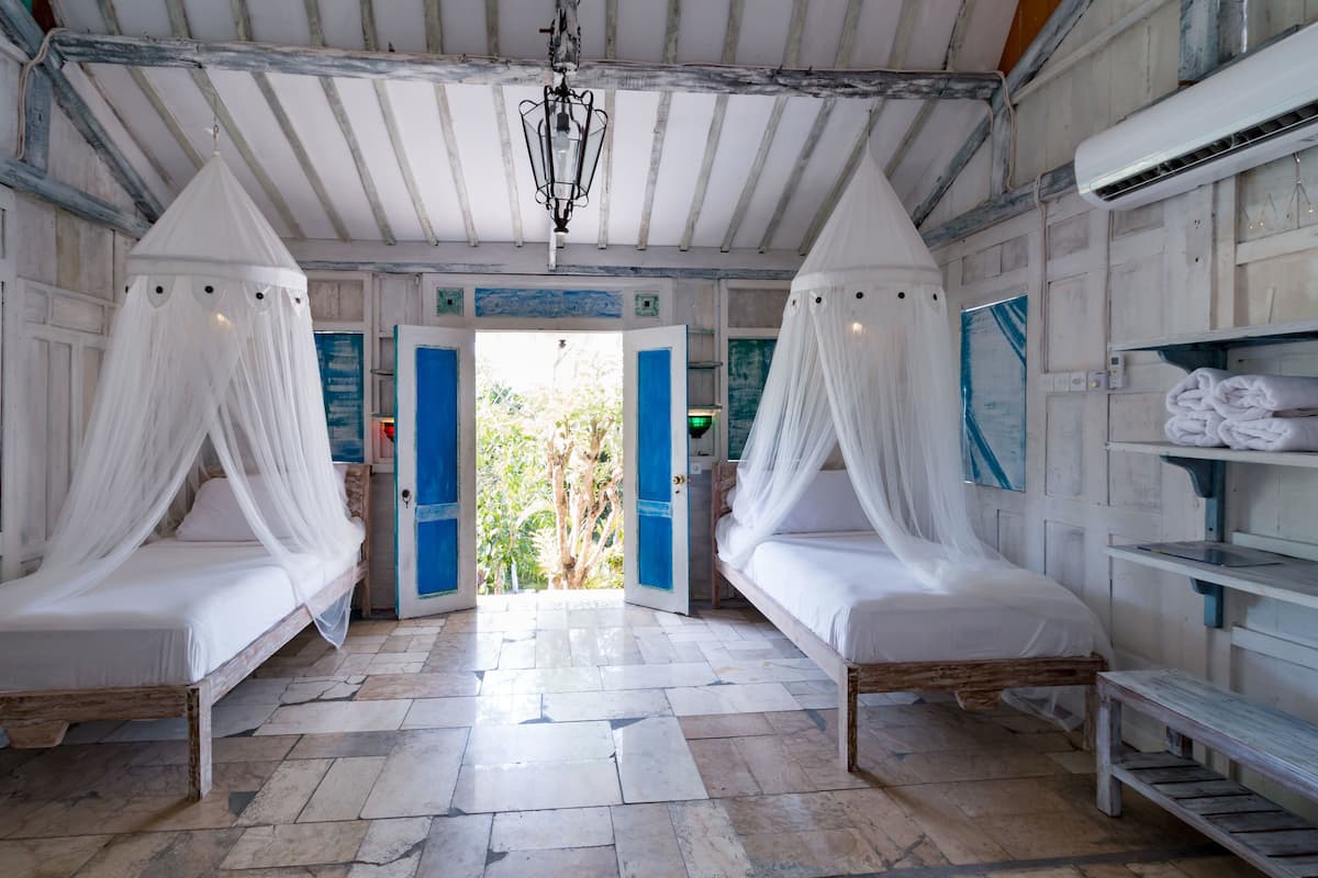 A room with two beds, each with a canopy, a wooden bench, and a view of greenery outside.