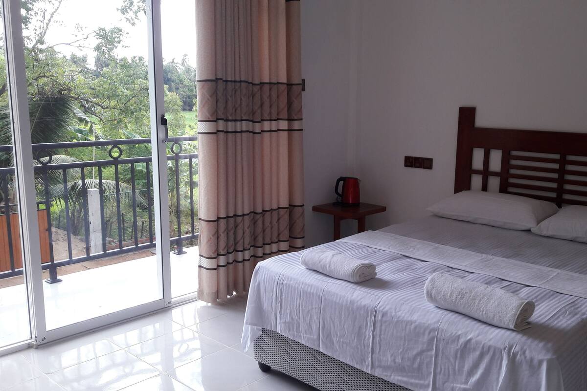 A hotel room with a large bed, a small table with a red kettle, and a view of greenery through the balcony doors.