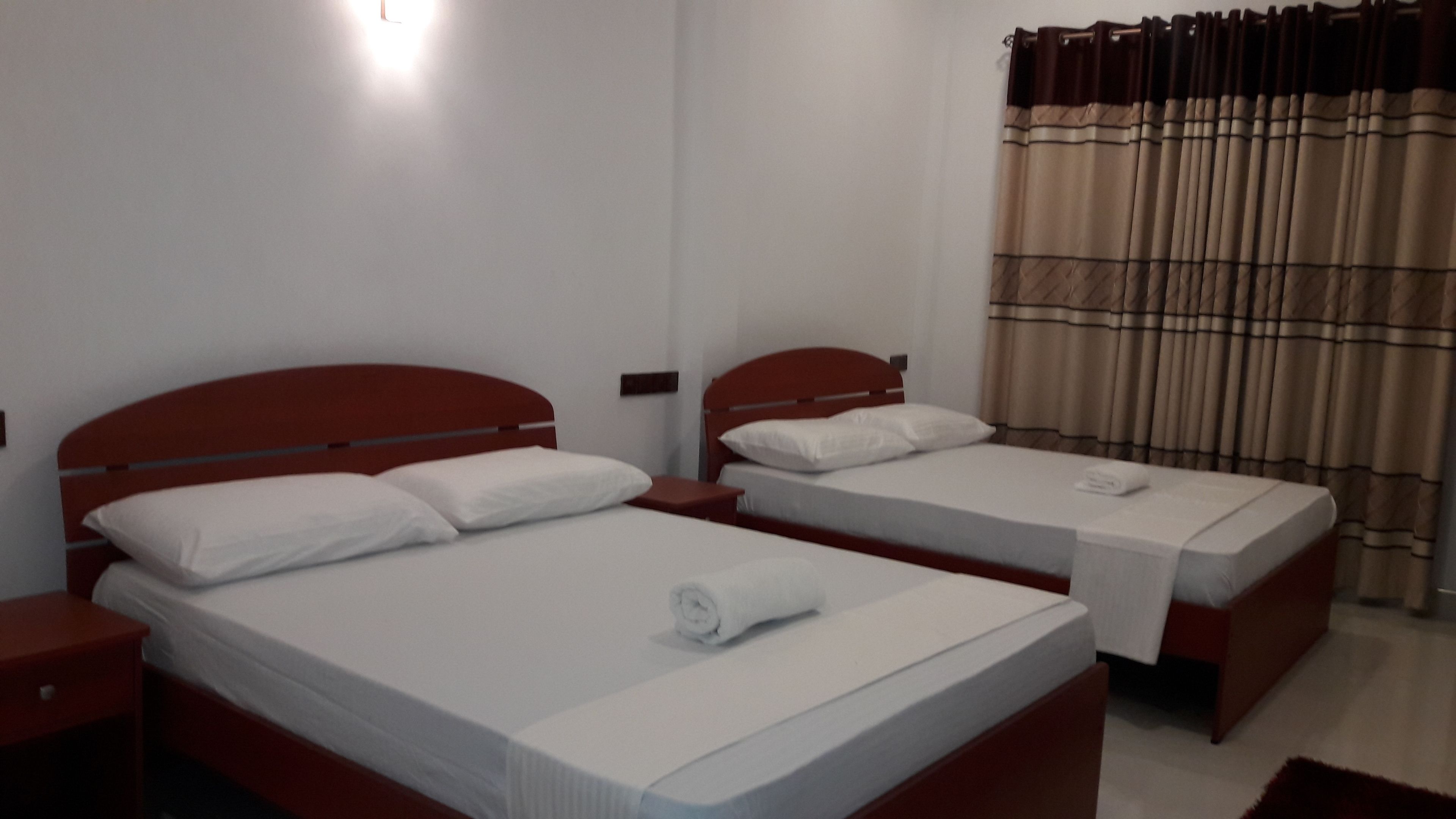 A hotel room with two single beds, each with a wooden headboard and white bedding. There is a small bedside table with a lamp on each bed. The room has a window with curtains.