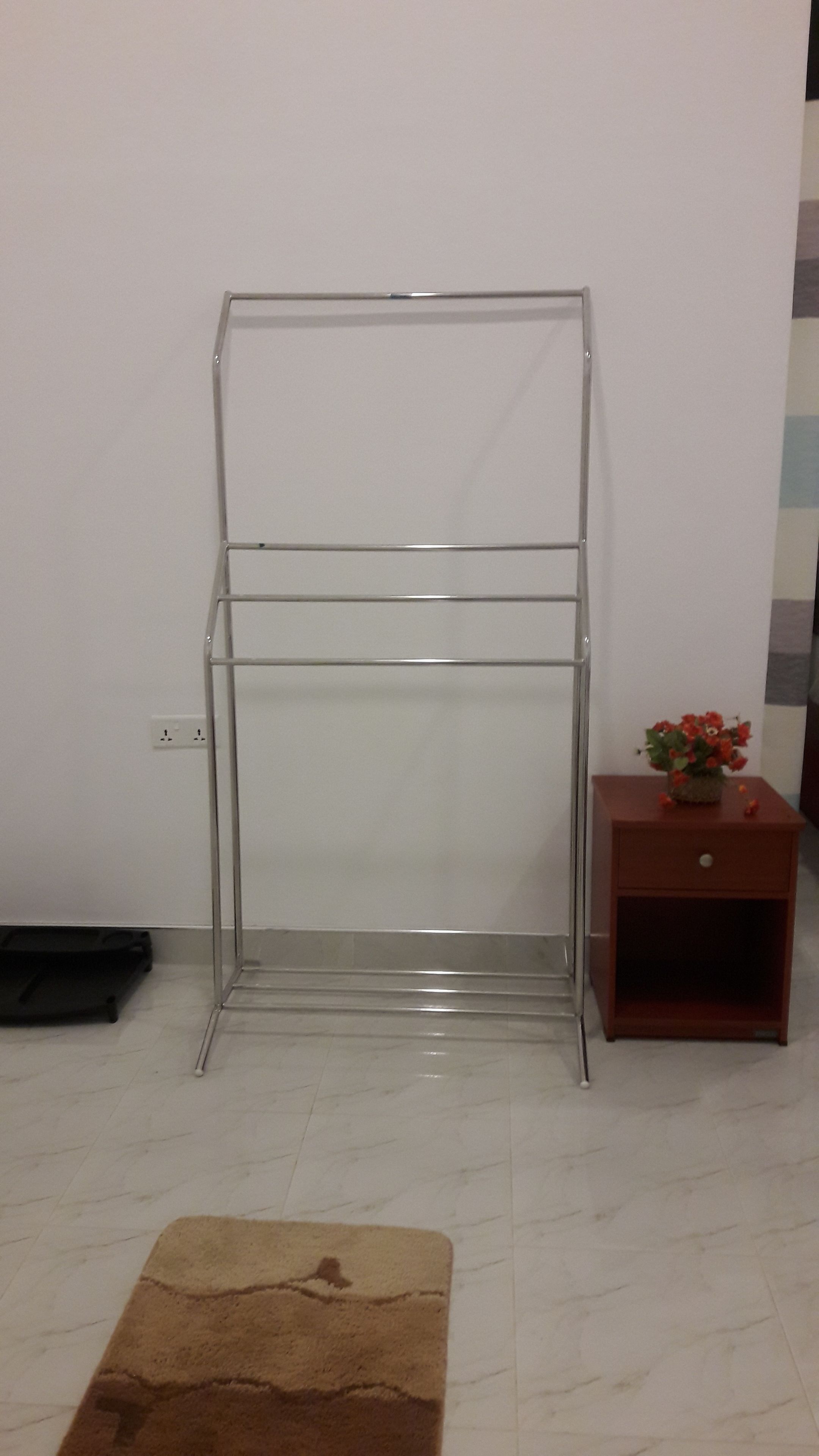 Una habitación con un tendedero de metal, una pequeña mesita de noche con un florero y una alfombra marrón en el suelo.