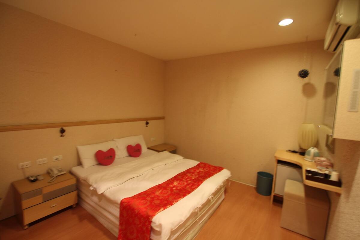 A hotel room with a bed, bedside table, nightstand, desk, chair, and a small trash bin.