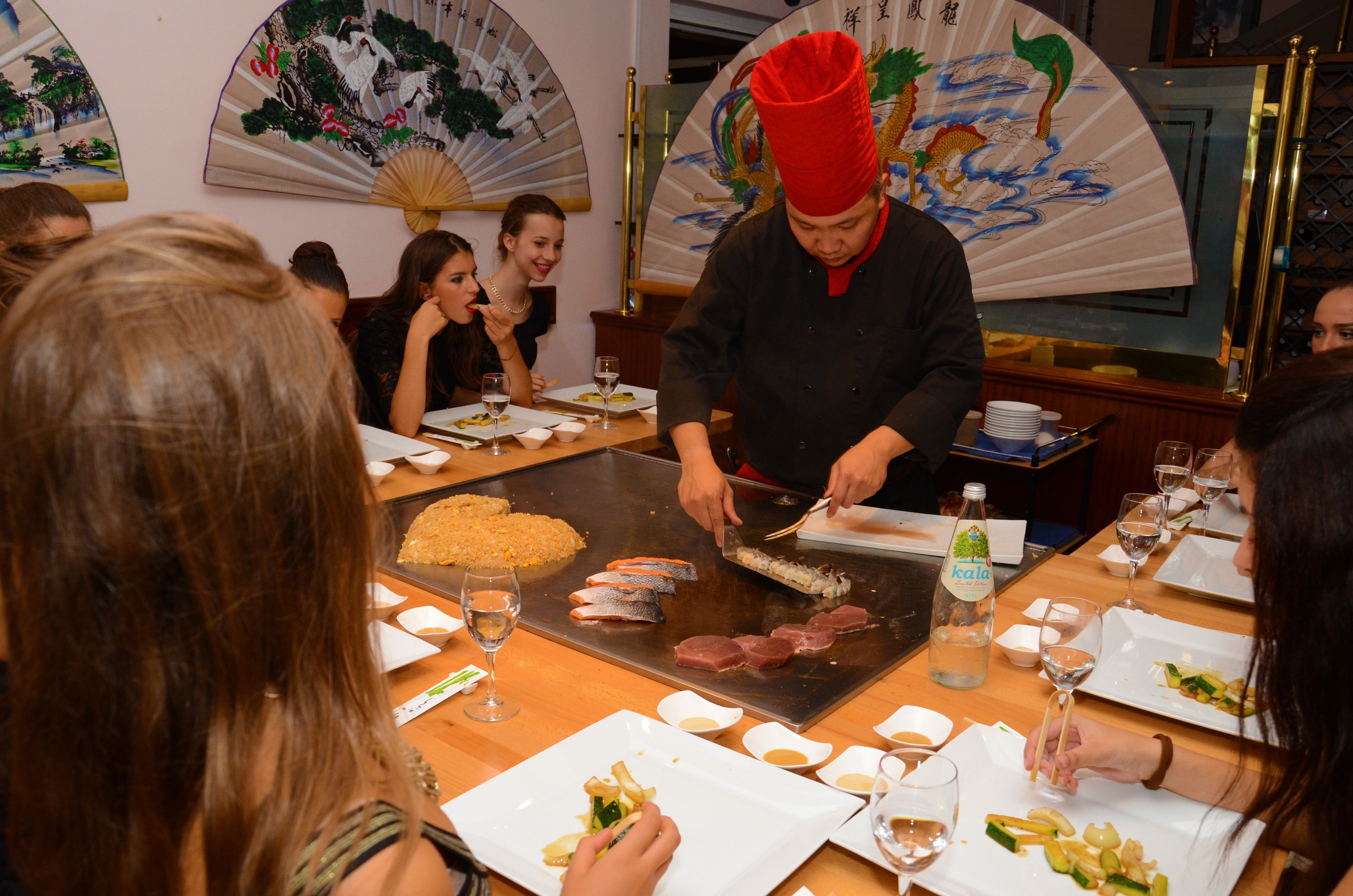 A dining area with a chef preparing food at a grill, guests seated at tables with plates and glasses, and traditional decor including fans and artwork.