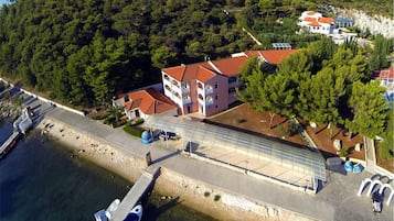 An aerial view of a coastal property with a large building, a dock with boats, and a forested hill in the background.