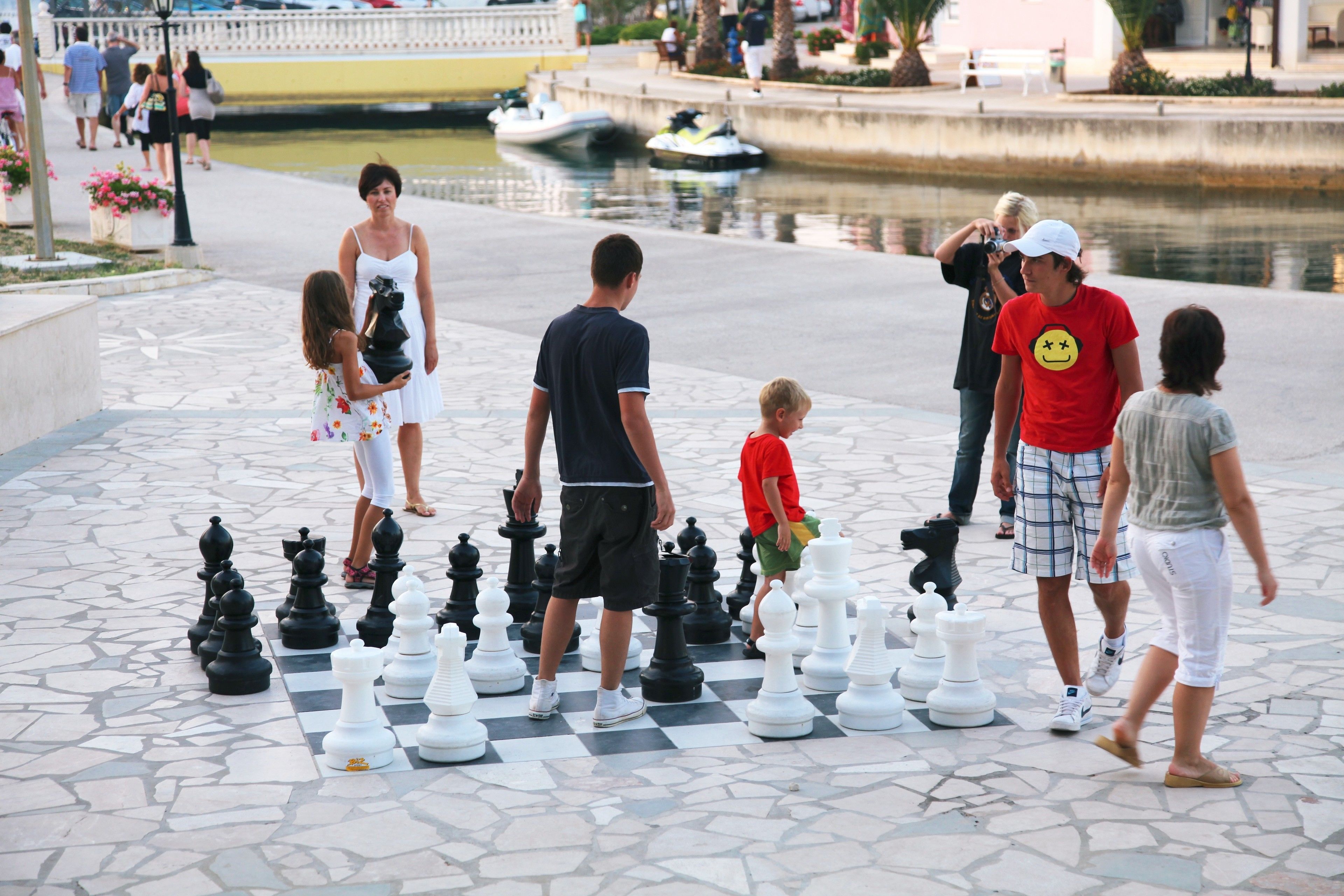 A chessboard with large pieces set up outdoors, surrounded by people.