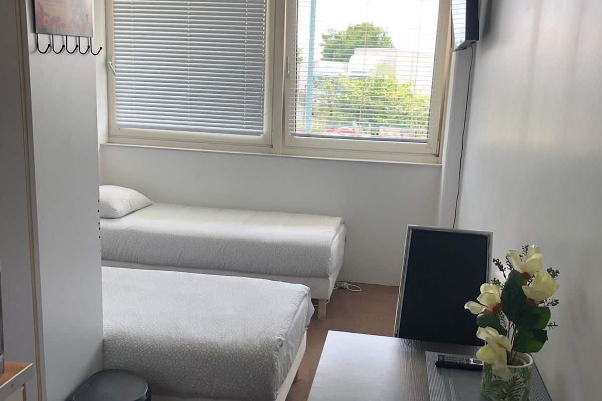 Une chambre de taille modeste, comprenant deux lits simples, un bureau avec un ordinateur et une plante en pot, une fenêtre avec des stores et une poubelle.
