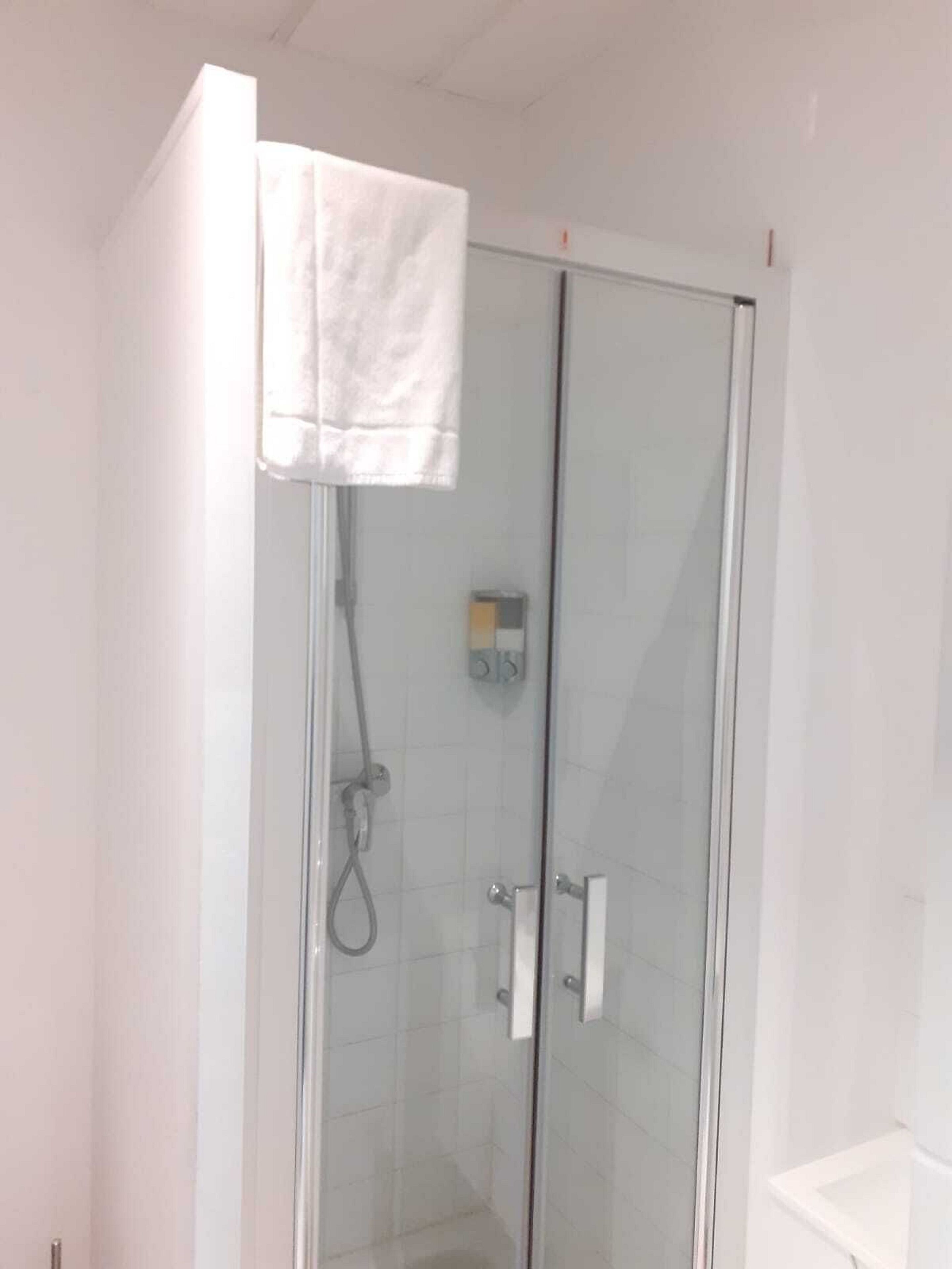 Una ducha con cerramiento de vidrio, un panel de control, una toalla blanca colgada en la puerta y un lavamanos blanco a la derecha.