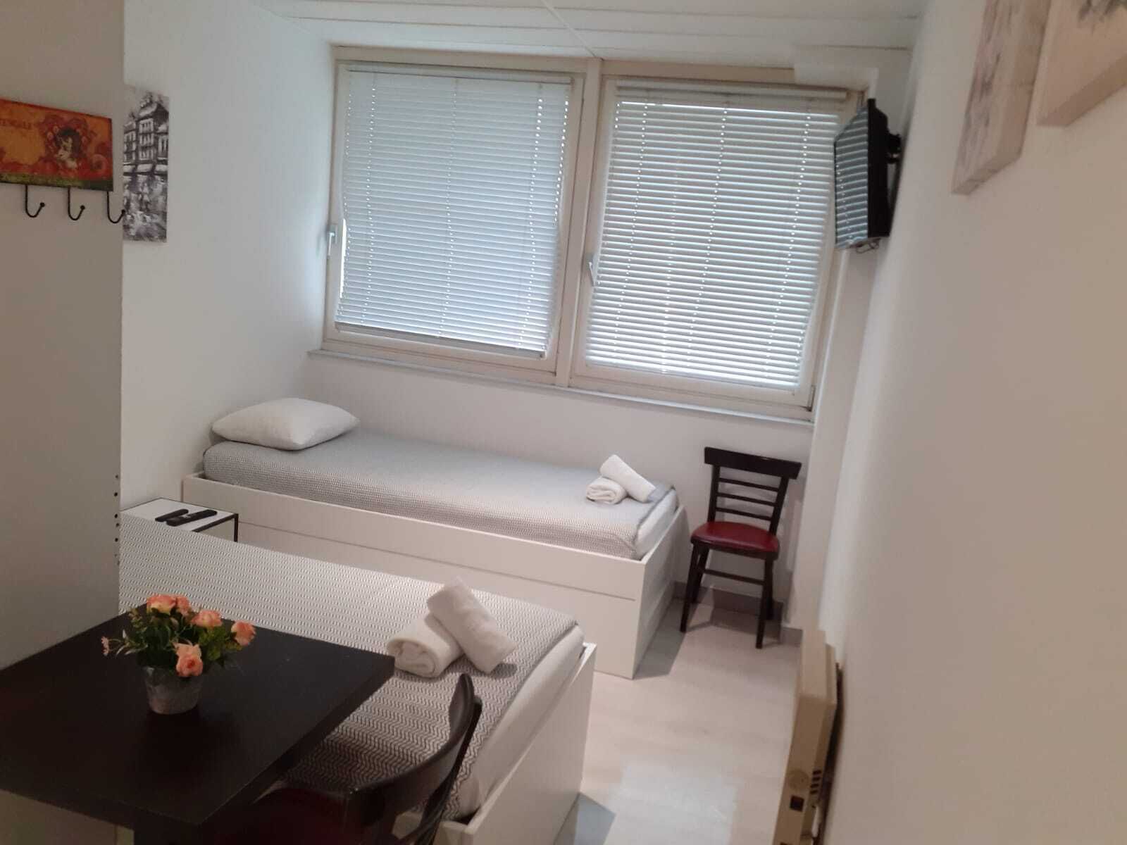 A small, compact room with a bed, a chair, a table, and a window with blinds.