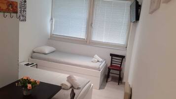 A small, compact room with a bed, a chair, a table, and a window with blinds.
