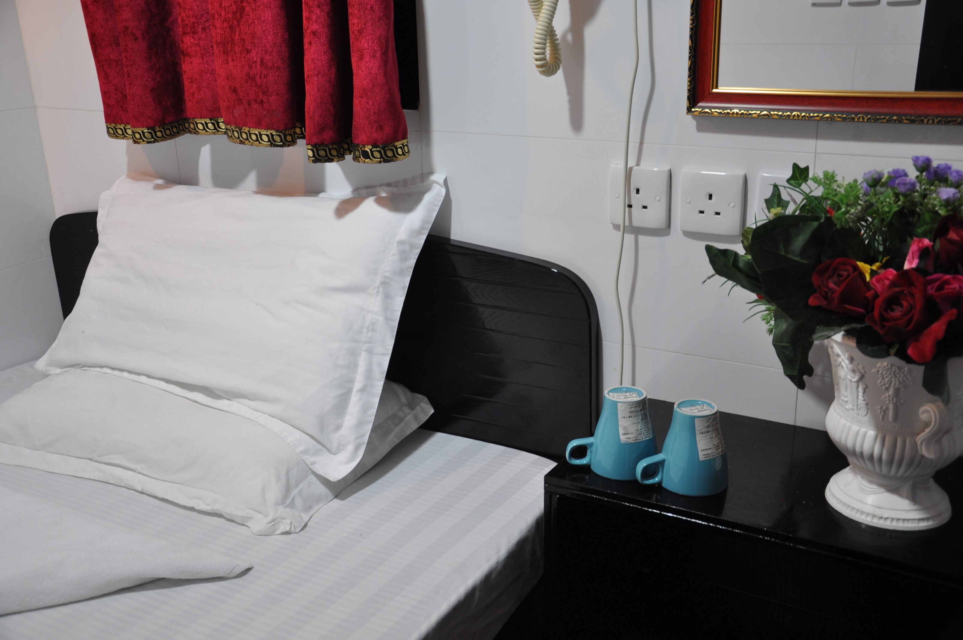 A neatly made bed with white linens, a black headboard, and a red curtain with a gold trim. There is a white vase with red roses on a black table beside the bed.