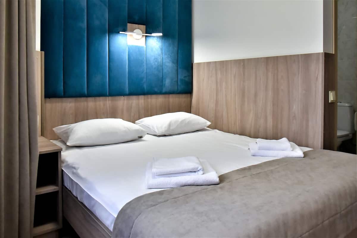 Un letto rifatto con lenzuola e asciugamani bianchi, una testiera in legno con un pannello blu decorativo e un comodino con una lampada.