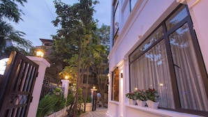 Un sendero estrecho que conduce a un edificio con fachada blanca, ventanas grandes y plantas en macetas en el alféizar.