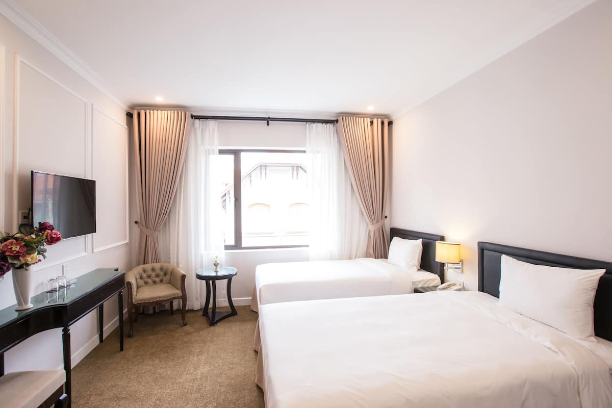 A hotel room with two beds, a TV, a desk with a vase of flowers, a chair, a small table, and a window with curtains.