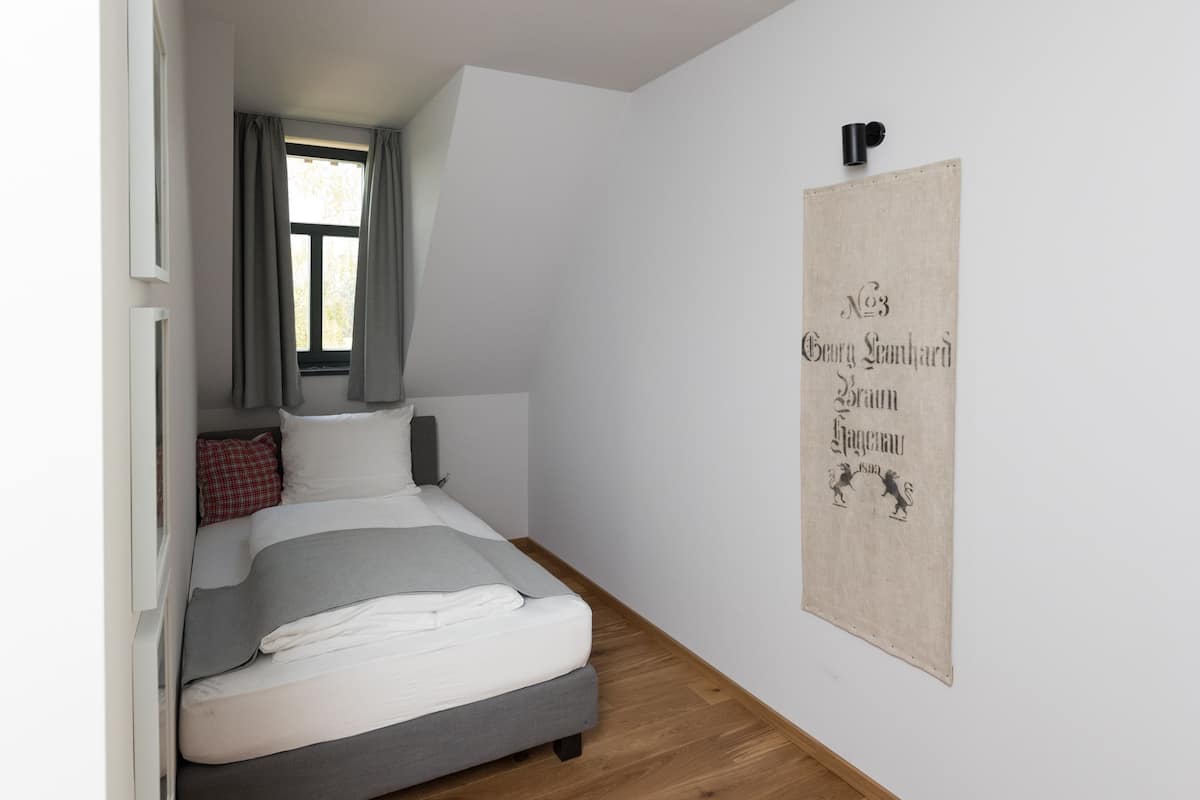 Une chambre à coucher avec un lit, une fenêtre avec des rideaux, un plancher en bois et un luminaire fixé au mur.