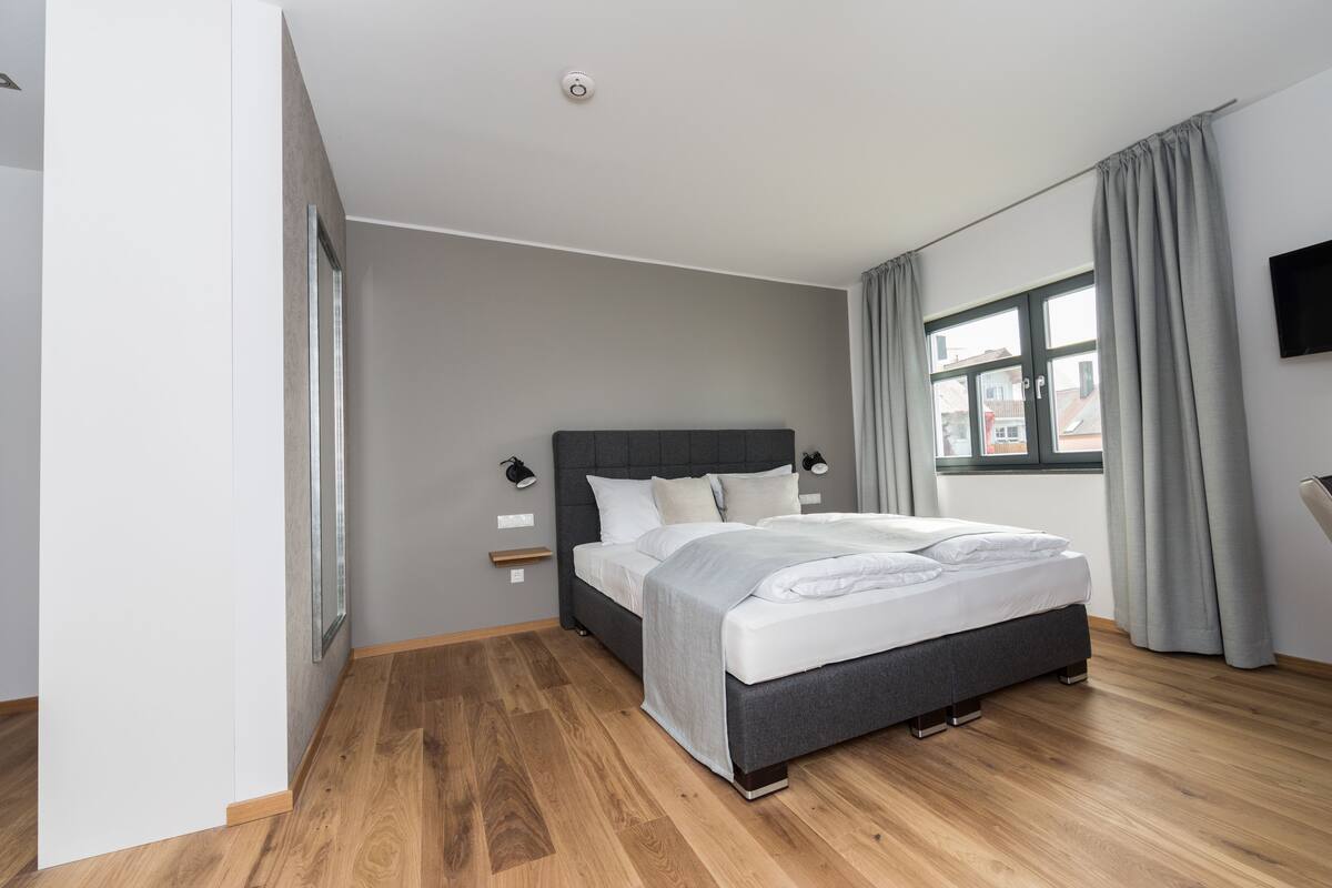 Une chambre à coucher moderne avec un grand lit, une table de chevet, une commode, une fenêtre avec des rideaux et un téléviseur fixé au mur.