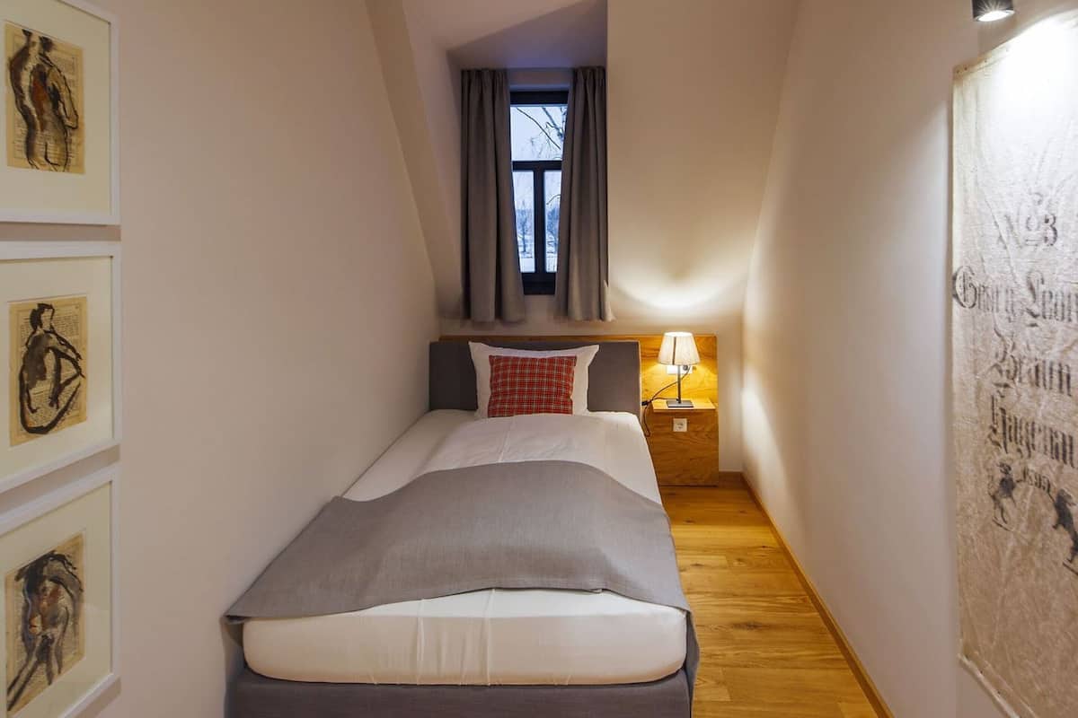 Une chambre d’hôtel étroite, avec un lit, une table de chevet, une fenêtre avec des rideaux et des œuvres d’art encadrées accrochées aux murs.
