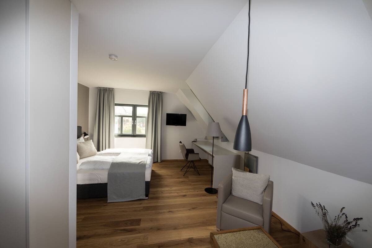 Une chambre d’hôtel moderne avec un plancher en bois, un lit recouvert d’une couverture grise, un bureau avec une chaise, une lampe et un téléviseur fixé au mur.