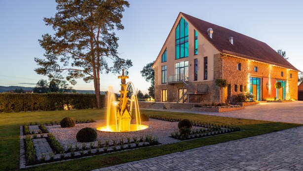 Een groot stenen gebouw met een opvallende fontein op de voorgrond, omringd door verzorgd groen en een heldere lucht.