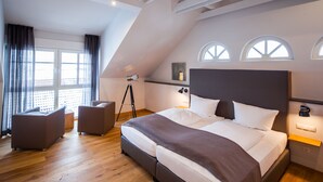 Een moderne slaapkamer met een groot bed, twee fauteuils, een driepoot lamp en uitzicht op de stad door grote ramen.
