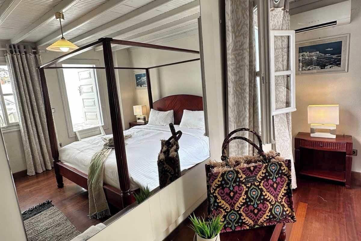 Quarto com cama de dossel, um espelho grande, mesa de cabeceira com abajur, uma bolsa decorativa e uma pequena planta em vaso.