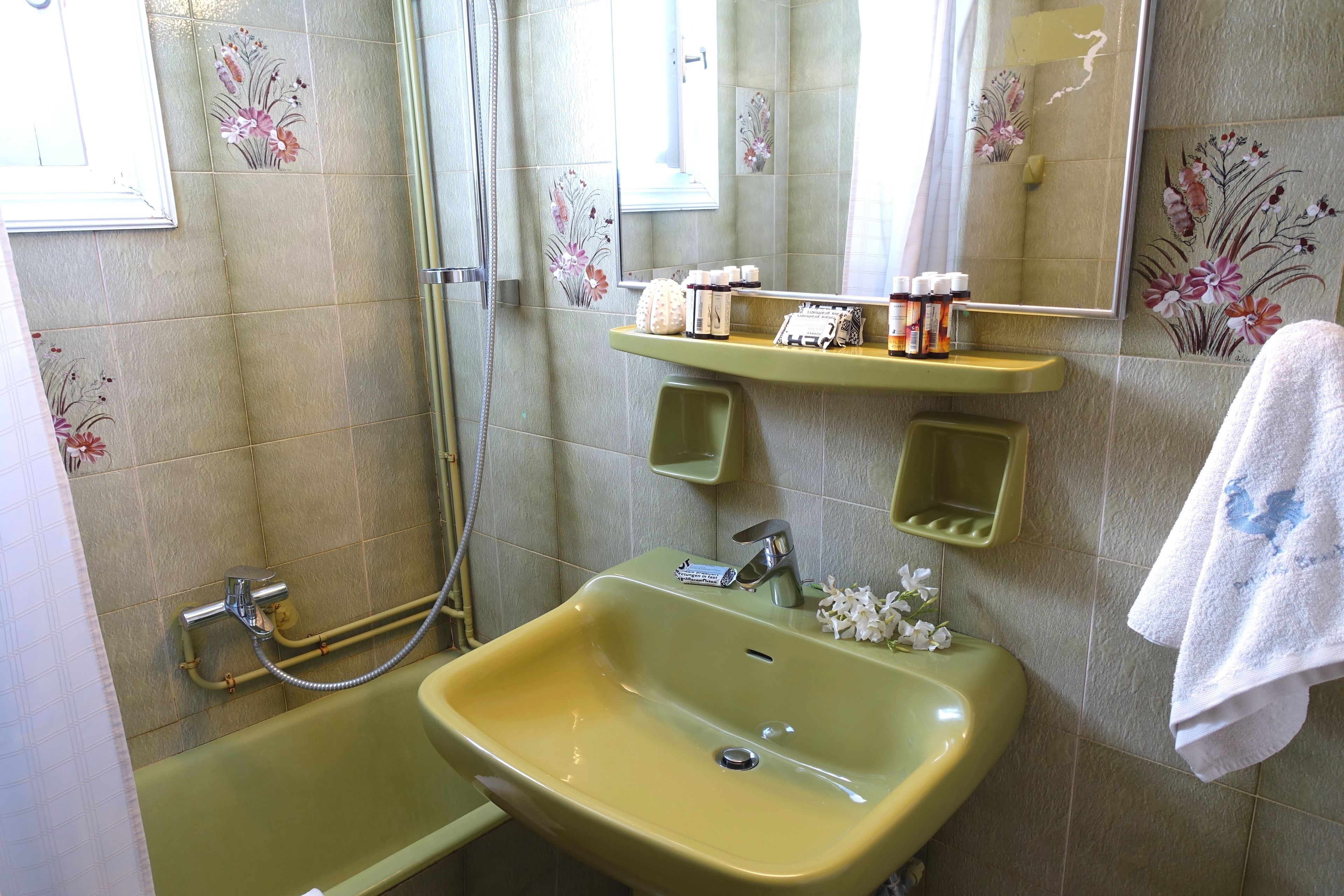 Une salle de bain avec un lavabo vert, un miroir et une douche. On trouve des articles de toilette sur le lavabo et une serviette est accrochée au mur.